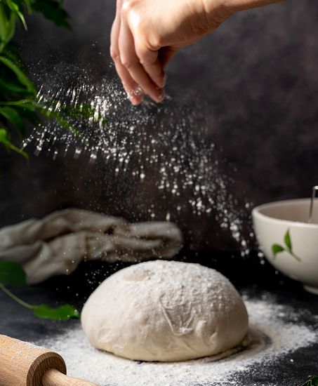 Person using their fingers to sift flour onto a piece of dough with a rolling pin and bowl visible in the shot